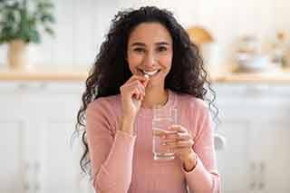 Woman in pink sweater holding glass of water in one hand and pill to her smile in the other